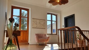 Une pièce avec un plancher en bois, une chaise rose, un vase vert contenant des fleurs séchées, une carte accrochée au mur et un escalier avec une rampe en bois.