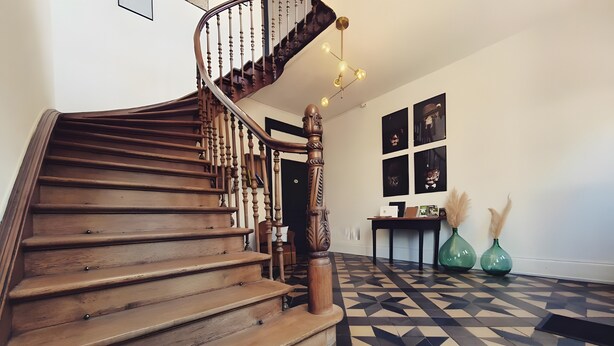 Un escalier en bois avec une rampe, un sol orné de motifs géométriques et un mur décoré d’œuvres d’art encadrées.