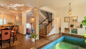 A spacious indoor swimming pool area with a staircase, a dining area with chairs, and a kitchen in the background. - Alaya Stays 3 BHK Casa Bella Vista  (Nerul)