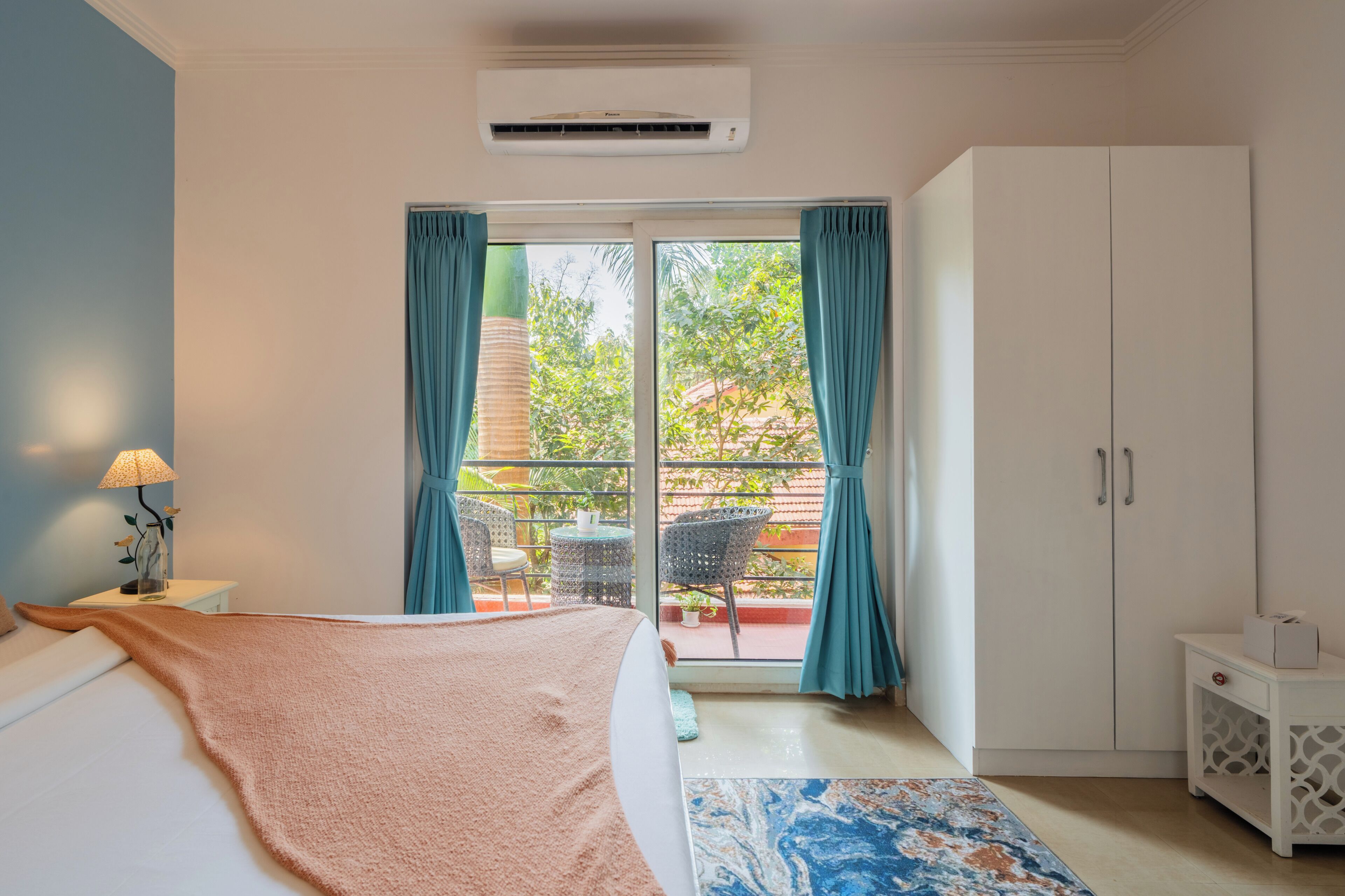 Un dormitorio con cama, mesita de noche, lámpara, ventana con cortinas, balcón con sillas y armario.