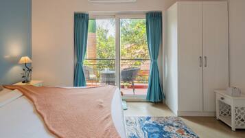 Un dormitorio con cama, mesita de noche, lámpara, ventana con cortinas, balcón con sillas y armario.