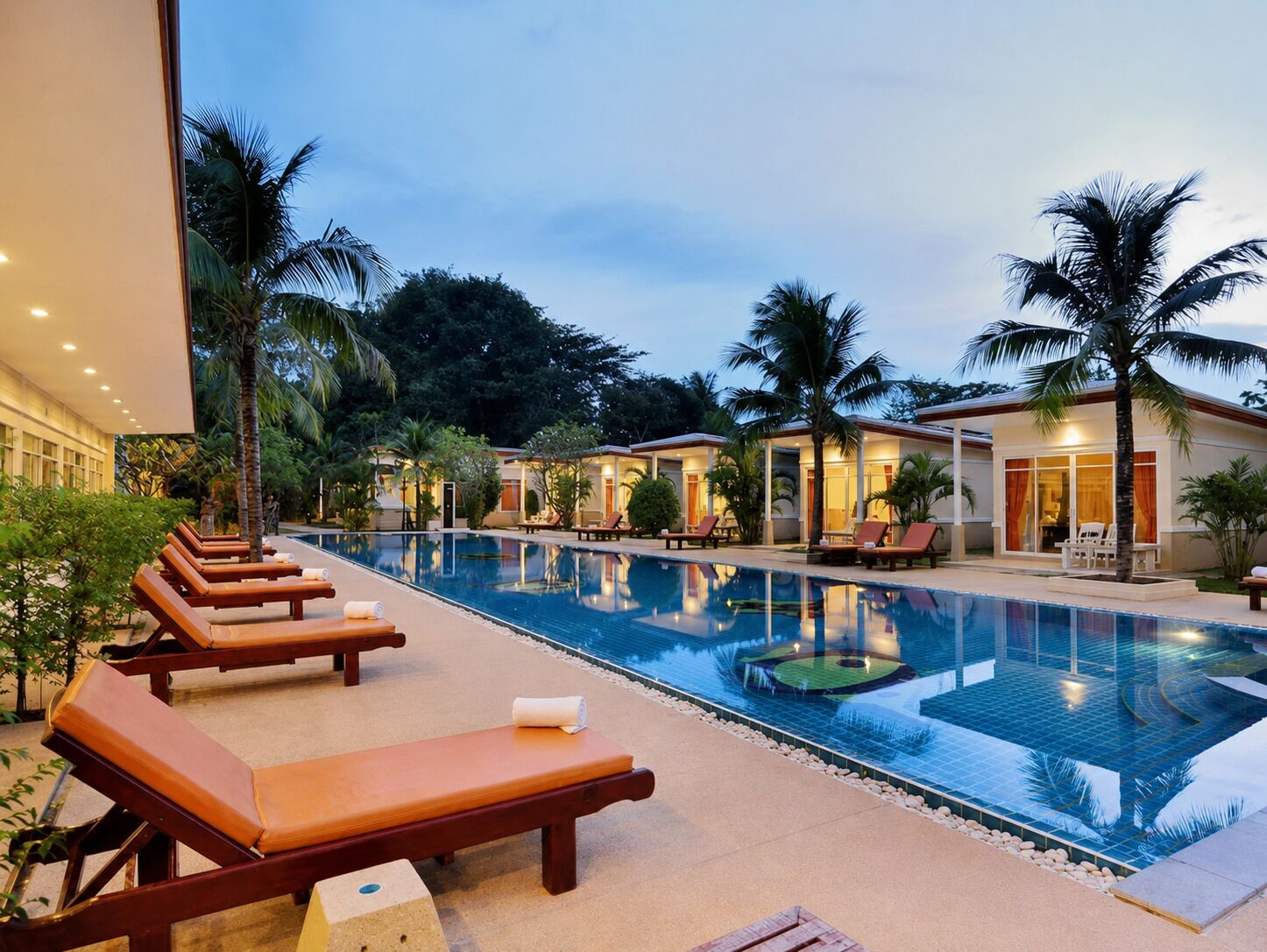 Un área de piscina con sillones de descanso y una piscina, rodeada de palmeras y edificios.