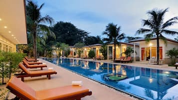 Un área de piscina con sillones de descanso y una piscina, rodeada de palmeras y edificios.