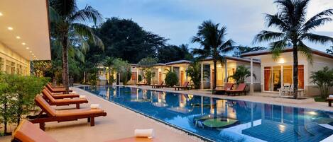 Un área de piscina con sillones de descanso y una piscina, rodeada de palmeras y edificios.