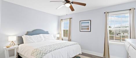 A bedroom with a bed, nightstands, a ceiling fan, a window with curtains, and a view of the outside.