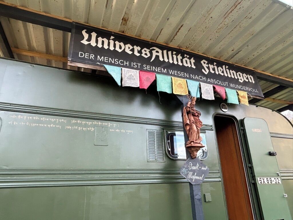 Ein alter Wohnmobilwagen mit einem Schild darüber, auf dem 'UniversAllität Stuttgart' steht, und einer Fahnenkette mit bunten Wimpeln.