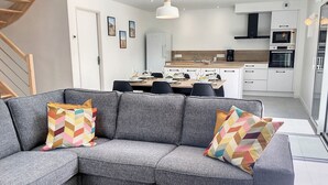 Un soggiorno moderno con un divano angolare grigio, una zona pranzo con tavolo in legno e una cucina con mobili e elettrodomestici bianchi.