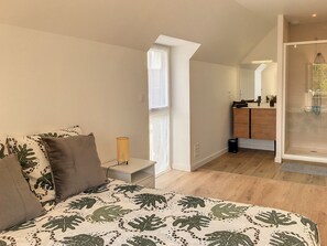 Ein Schlafzimmer mit Bett, Nachttisch, Lampe, Fenster und einem Badezimmer mit Dusche und Spiegel.