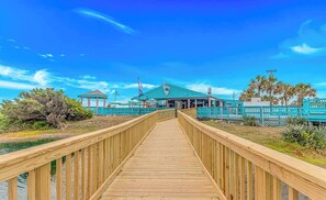 A wooden boardwalk leading to a building with a green roof and a flag. - Perfect Paradise (Myrtle Beach)