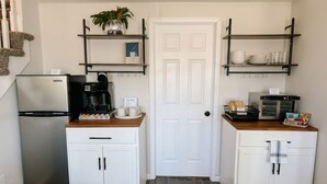 Une cuisine compacte avec des meubles blancs, un réfrigérateur en acier inoxydable, une machine à café et un grille-pain. Il y a deux étagères au-dessus des meubles : l’une est ornée d’une plante, l’autre contient de la vaisselle et une cafetière.
