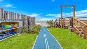 A beachside walkway leading to a wooden deck with a 'Welcome' sign, a beach umbrella, and a wooden fence with beach toys.