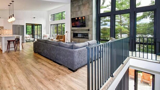 A spacious living area with a large gray sectional sofa, a kitchen with white cabinetry, and a stone fireplace. - Serene 6-bedroom Retreat in Tory Hill With Lake Views (North Kawartha)