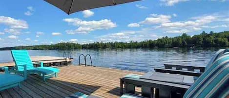A wooden deck with lounge chairs and a table, overlooking a calm lake with a forested shoreline.