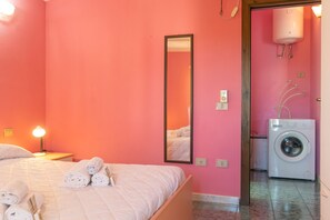 Ein rosafarbenes Zimmer mit einem Bett, einem Spiegel, einem Nachttisch mit Lampe und einer Waschmaschine.