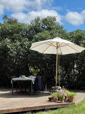 Une terrasse extérieure avec une table dressée pour le repas, entourée de plantes en pot et agrémentée d’un parasol.