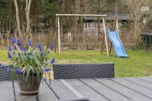 Une aire de jeux avec un toboggan et des balançoires, entourée d’arbres et d’une clôture.