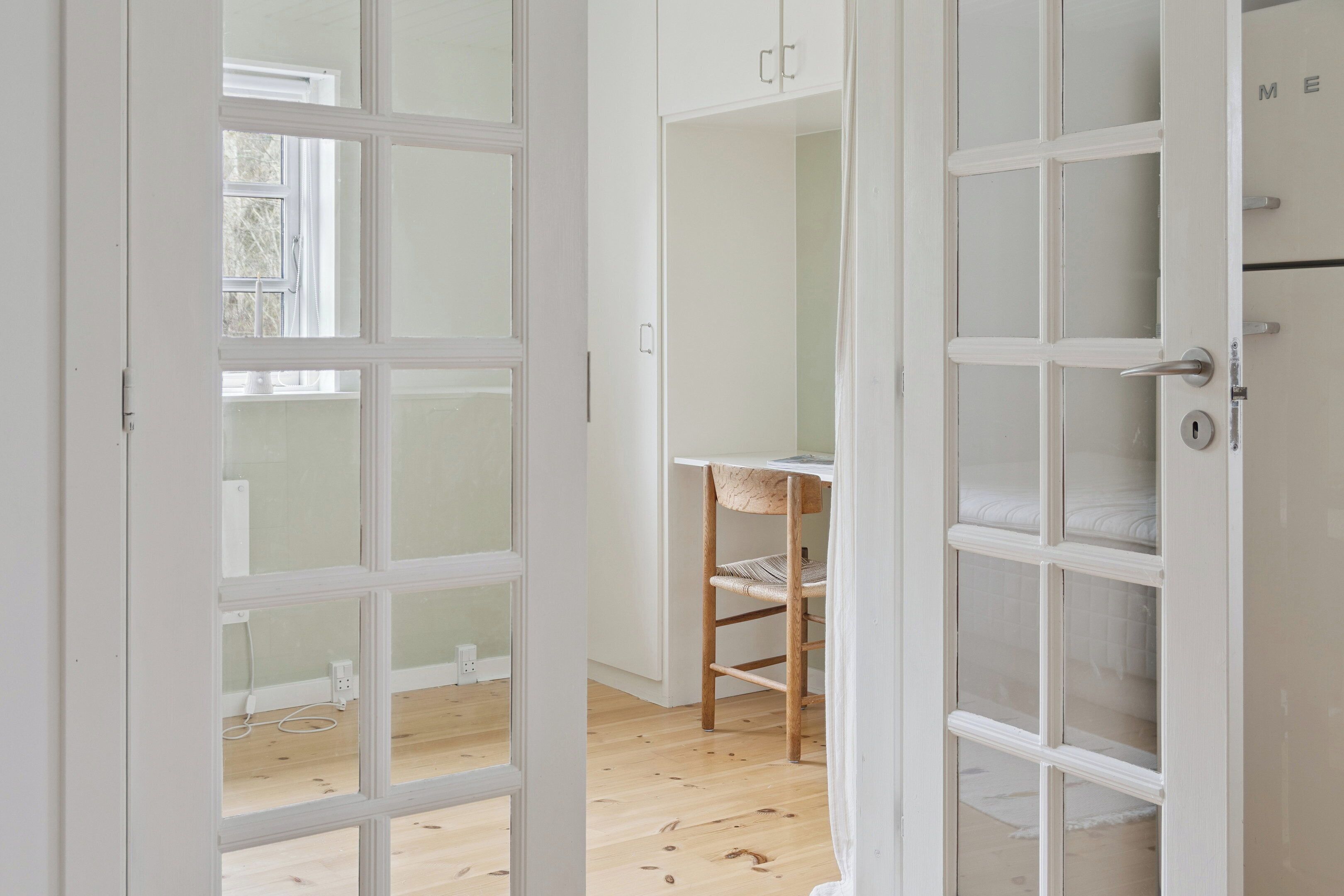 Une porte ouverte laisse entrevoir une pièce dotée d’un plancher en bois, d’une chaise et d’une armoire blanche avec des tiroirs.