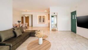 A modern living room with a sectional sofa, a coffee table with dried flowers, a flat-screen TV, and a dining area in the background. - Lithea Seaview Villa & Estate (Chania)