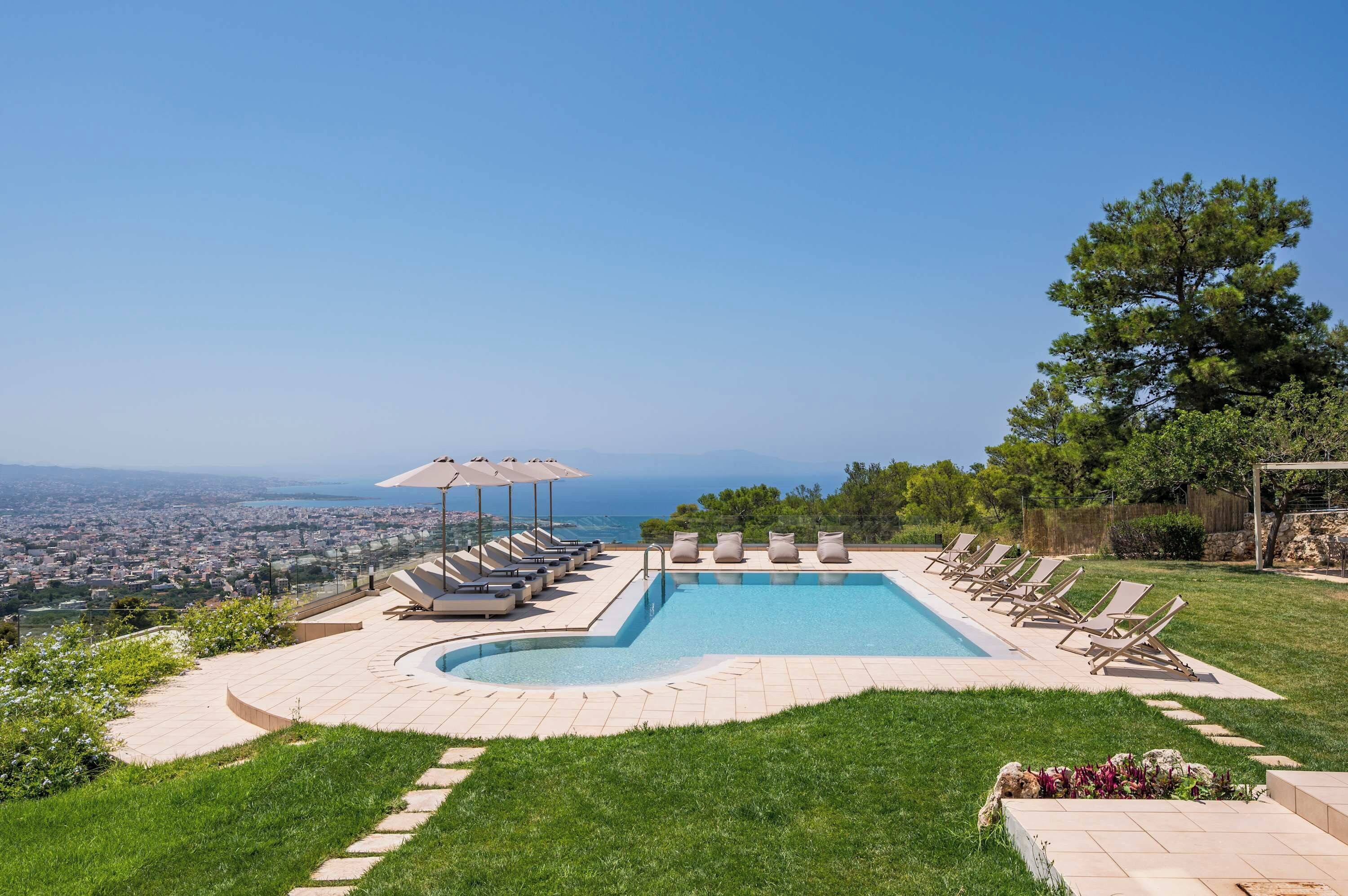 Ein Swimmingpool mit Liegestühlen und Sonnenschirmen, umgeben von einer gepflegten Rasenfläche und Blick auf eine Stadtlandschaft.