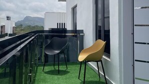 Un balcón con barandilla de vidrio, dos sillas, una puerta y vistas a montañas.