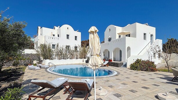 A pool area with lounge chairs and an umbrella, surrounded by white buildings with arches. - Villa Gala (Kamari)