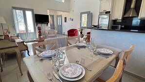 A dining area with a table set for four, a kitchen in the background, and a living room with a TV. - Villa Gala (Kamari)
