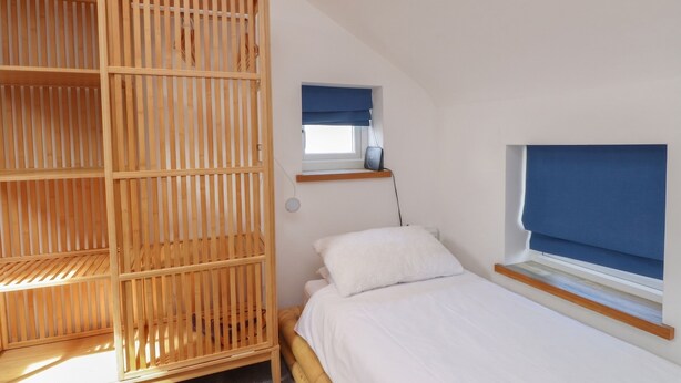 A bedroom with a wooden wardrobe, a bed with white bedding, a window with a blue blind, and a small shelf above the bed. - Kiloran (Rhosneigr)