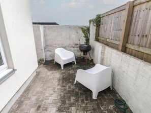 A small outdoor patio with two white plastic chairs, a barbecue grill, and a hose on the ground. - Kiloran (Rhosneigr)