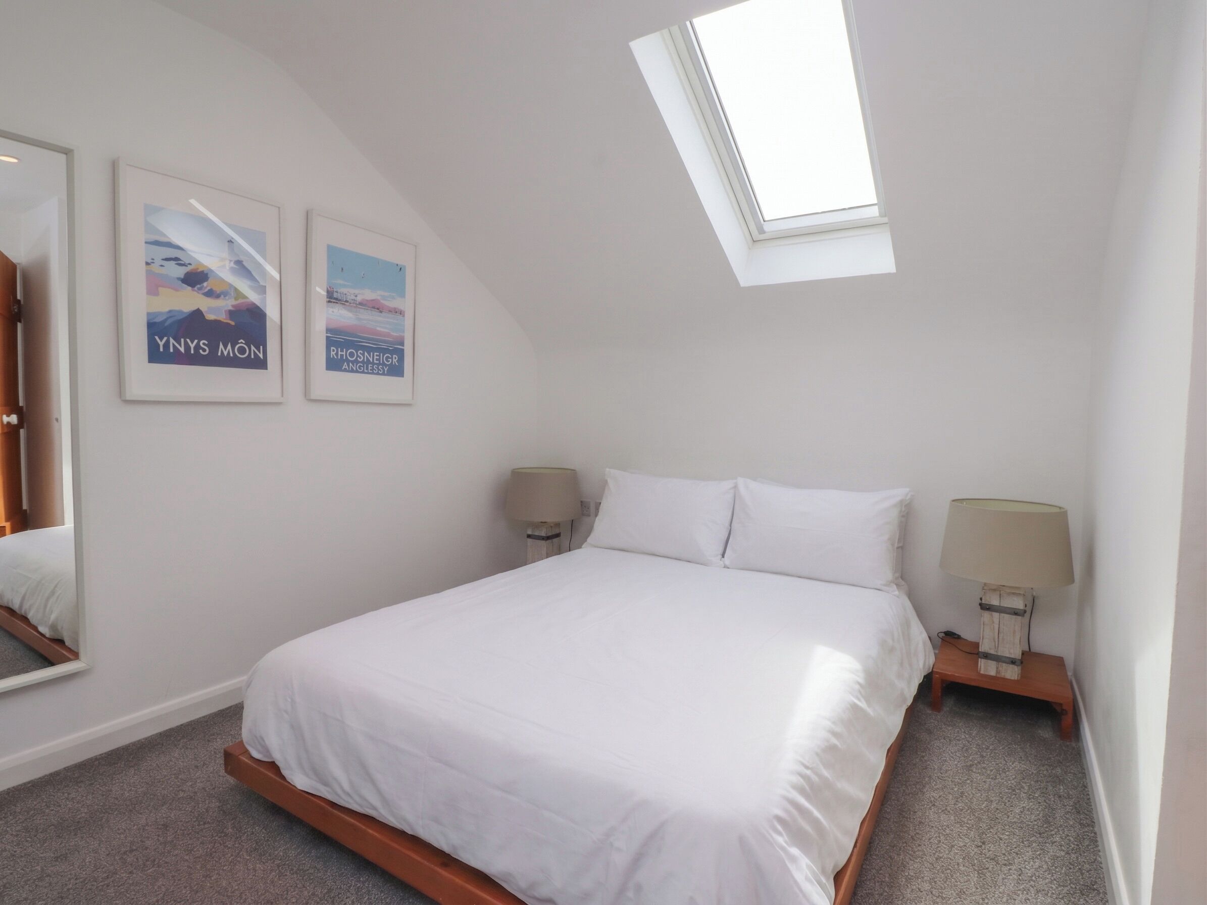 A bedroom with a skylight, a bed with white bedding, two bedside tables with lamps, and two framed pictures on the wall.