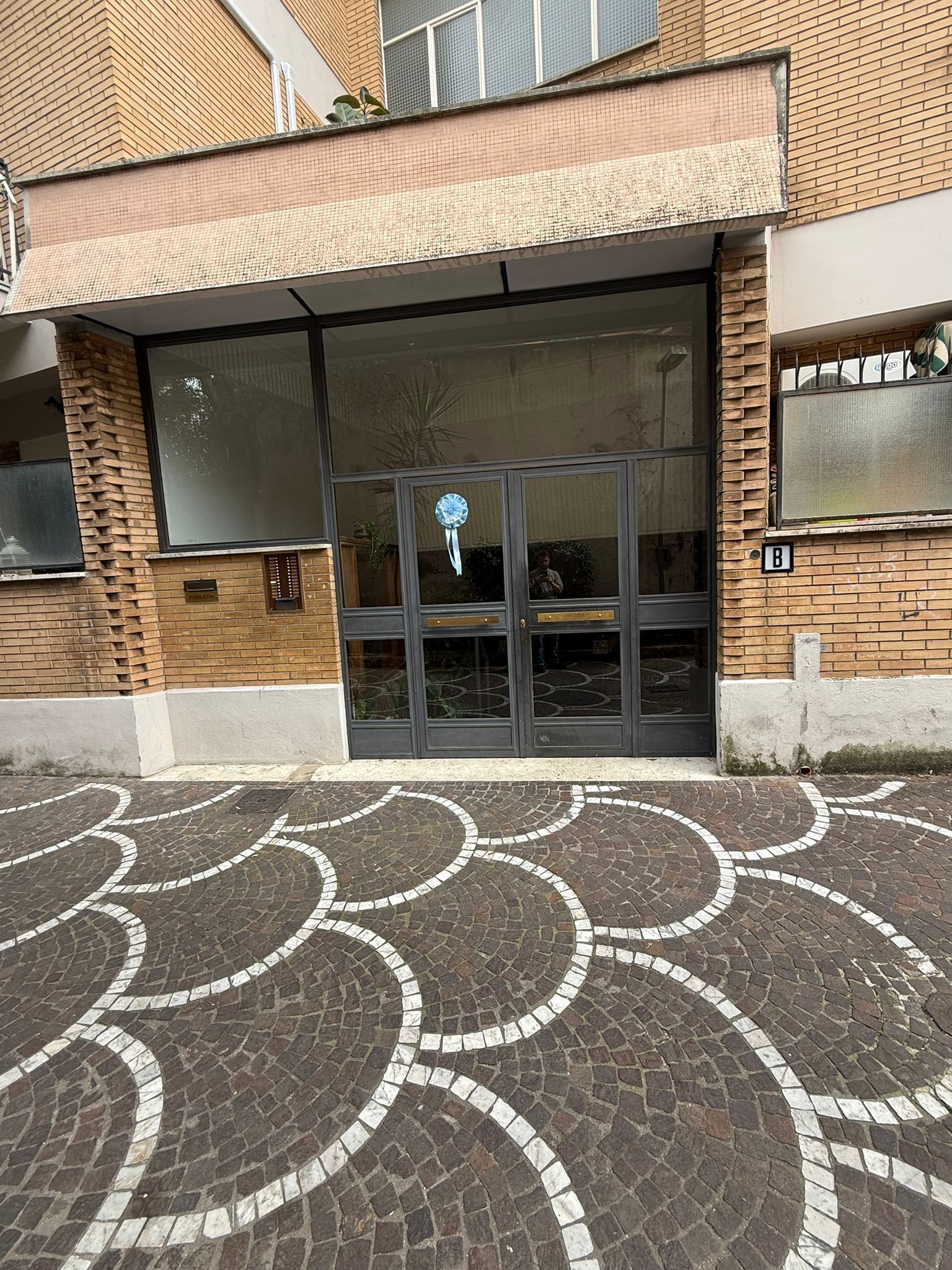 A building entrance with a glass door, a tiled pavement, and a brick facade.