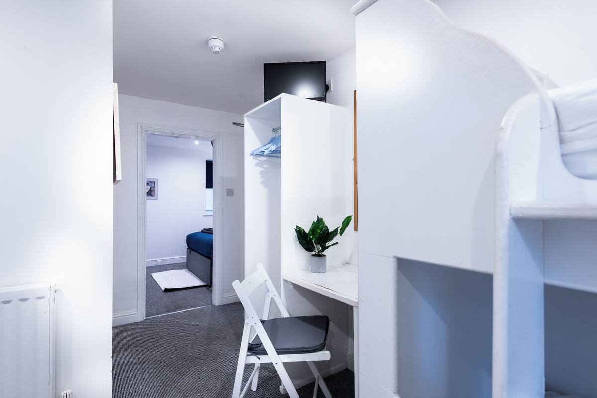 A modern room with a white desk, a chair, a small plant, and a bed in the background.
