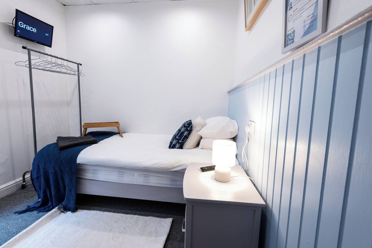 A bedroom with a bed, bedside table, lamp, TV, and a wall-mounted drying rack.