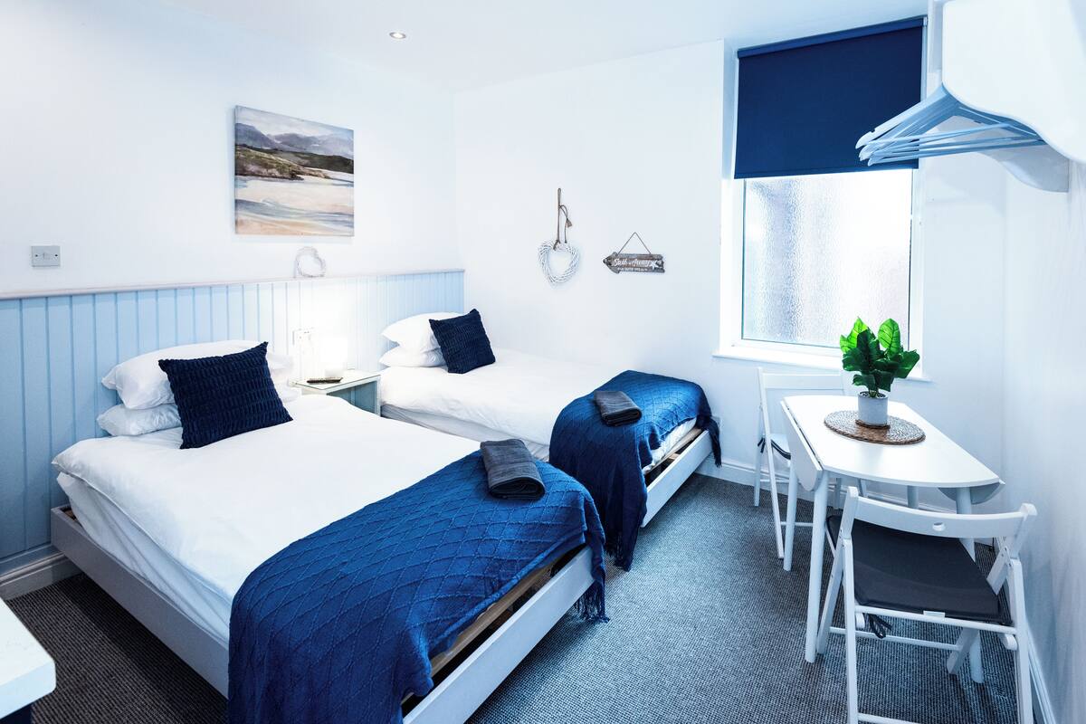 A hotel room with two beds, a small table, a chair, a window with a view, and a wall-mounted clock.