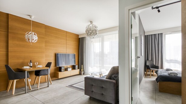 Quarto de hotel moderno com parede de detalhes em madeira, área de refeições com mesa e cadeiras, sofá, televisão e cama.