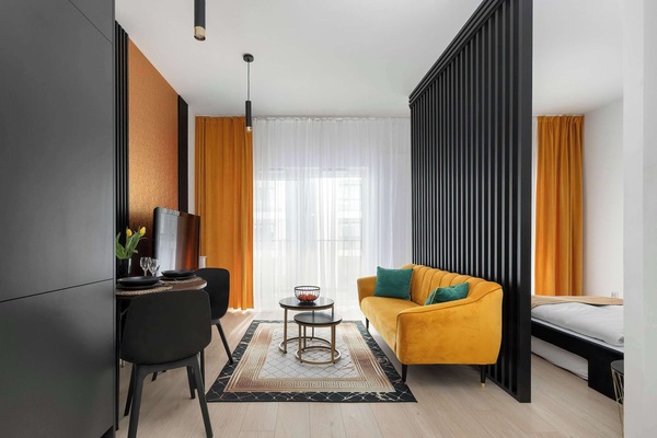A bright living room with a mustard-yellow sofa, a TV unit, and large windows that let in plenty of natural light. A small dining area and modern decor add to the cozy yet stylish atmosphere.