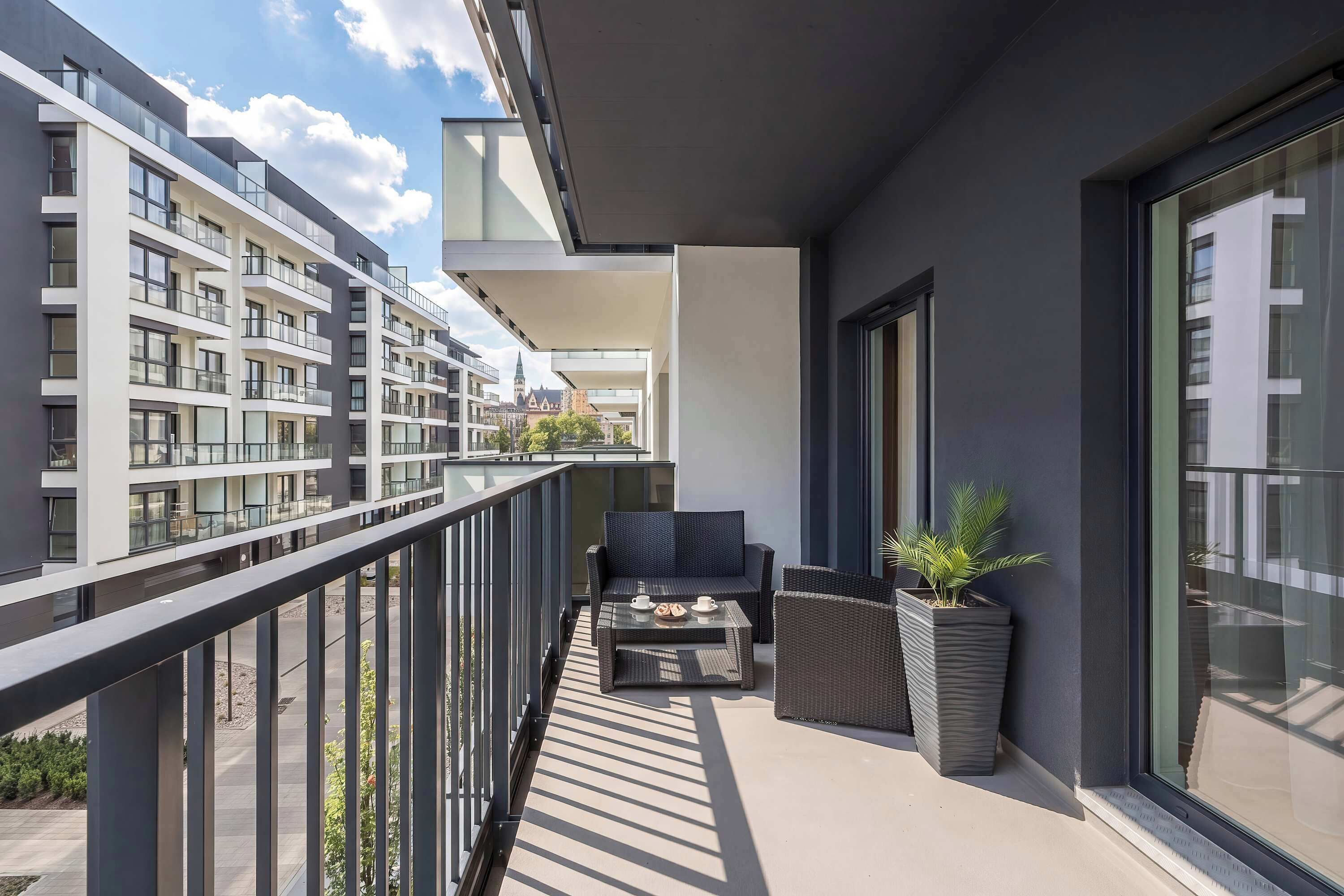 A long balcony with outdoor seating and a view of neighboring buildings. Potted plants add a touch of greenery.