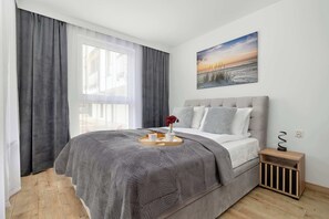 Ett sovrum med en säng, ett sängbord, en liten avlastningsyta, ett fönster med gardiner och en tavla som hänger på väggen.