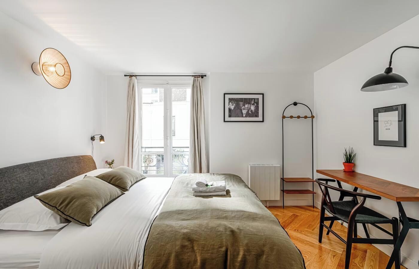A bedroom with a bed, bedside table, desk, chair, window, curtains, and wall-mounted lamp.