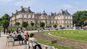 Ein großes, historisches Gebäude mit klassischer Architektur, umgeben von einem gepflegten Park mit einer kreisrunden Wiese und Sitzgelegenheiten.