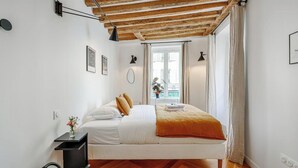 Ein Schlafzimmer mit Holzboden, einem Bett mit weißer Bettwäsche und orangefarbenen Kissen, einem Nachttisch mit Blumenvase und einem Fenster mit Blick auf Gebäude.