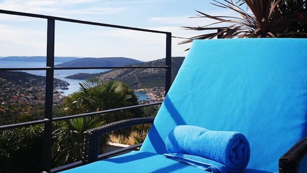 Een balkon met een blauwe zitting, een opgerolde handdoek en uitzicht op een kustlandschap.
