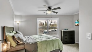 A bedroom with a bed, a ceiling fan, a window with a view of a parking lot, a dresser, and a lamp.
