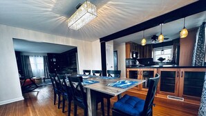 A dining area with a wooden table and chairs, a chandelier, and a kitchen in the background.