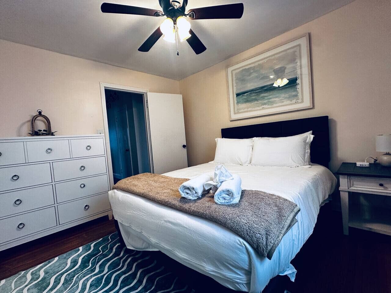 Een hotelkamer met een bed, een plafondventilator, een dressoir, een nachtkastje en een schilderij aan de muur.