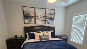 Ein Schlafzimmer mit Bett, Nachttisch, Schminktisch und Deckenventilator. An der Wand befindet sich ein Kunstwerk.