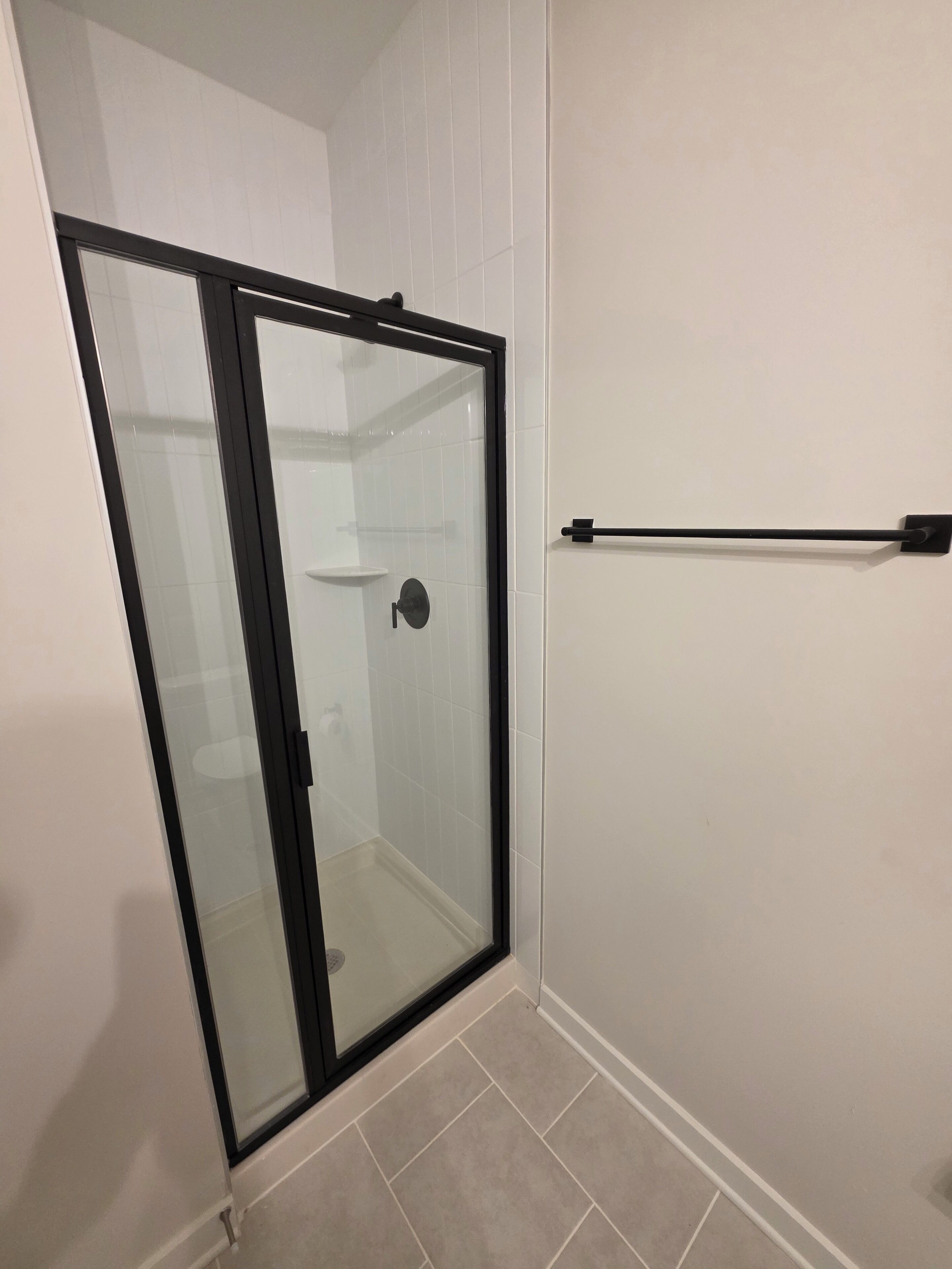 A glass-enclosed shower with a black frame, a white tiled wall, and a black towel rack.
