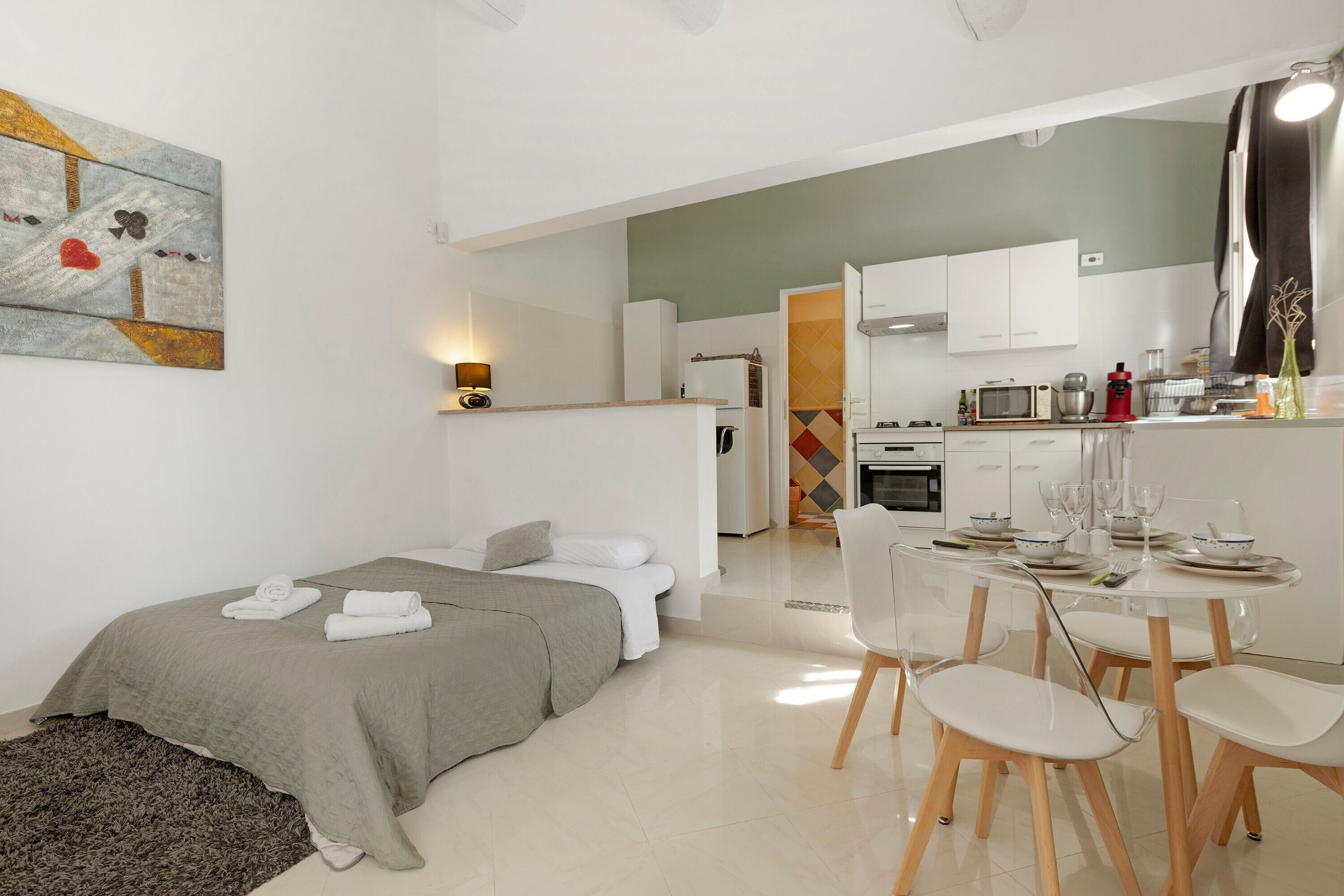 Ein modernes, helles Zimmer mit einem Bett, einem Esstisch mit Stühlen, einer Küchenzeile und einem Bild an der Wand.