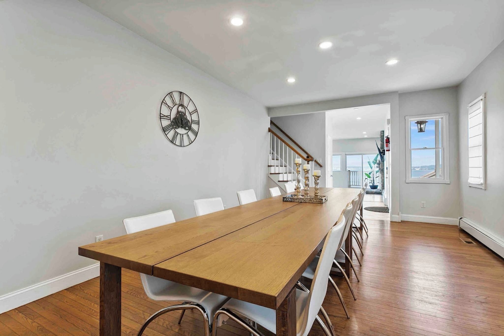 Uma sala de jantar com uma mesa de madeira comprida, cadeiras brancas, um relógio de parede e uma escada que leva a outro cômodo.