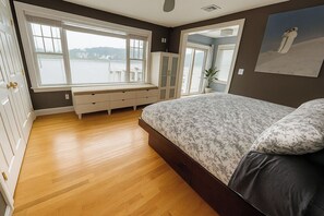 Une chambre avec un grand lit, un plancher en bois, une fenêtre donnant sur un plan d’eau, une armoire et une porte menant à une autre pièce.