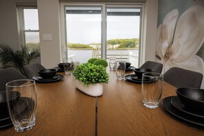 Mesa de jantar com tigelas pretas, copos e um vaso de planta, com vista para um lago e edifícios.